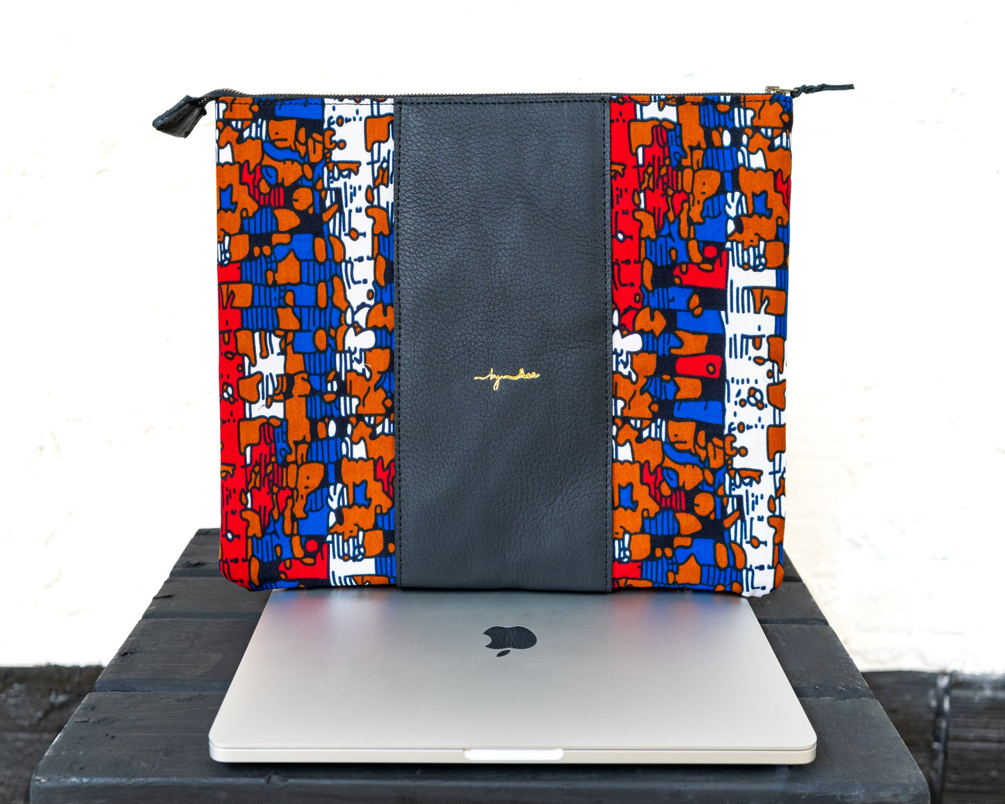 A multicolored laptop bag with a geometric pattern and a solid black panel, featuring a laptop partially inserted into it, displayed on a dark surface.